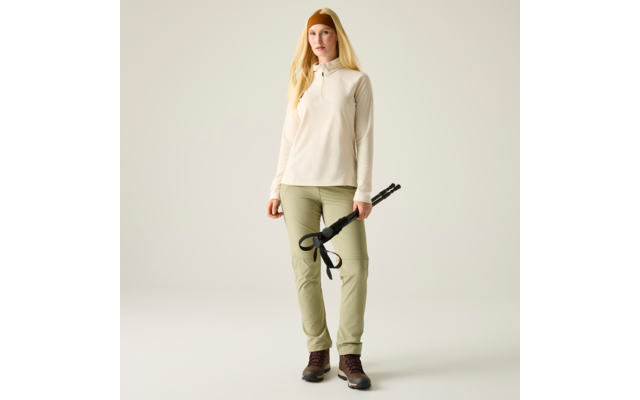 Frau mit langen blonden Haaren, beigem Pullover, olivgrüner Hose, braunen Wanderschuhen und orangem Stirnband, hält zusammengeklappte Wanderstöcke vor hellem einfarbigem Hintergrund