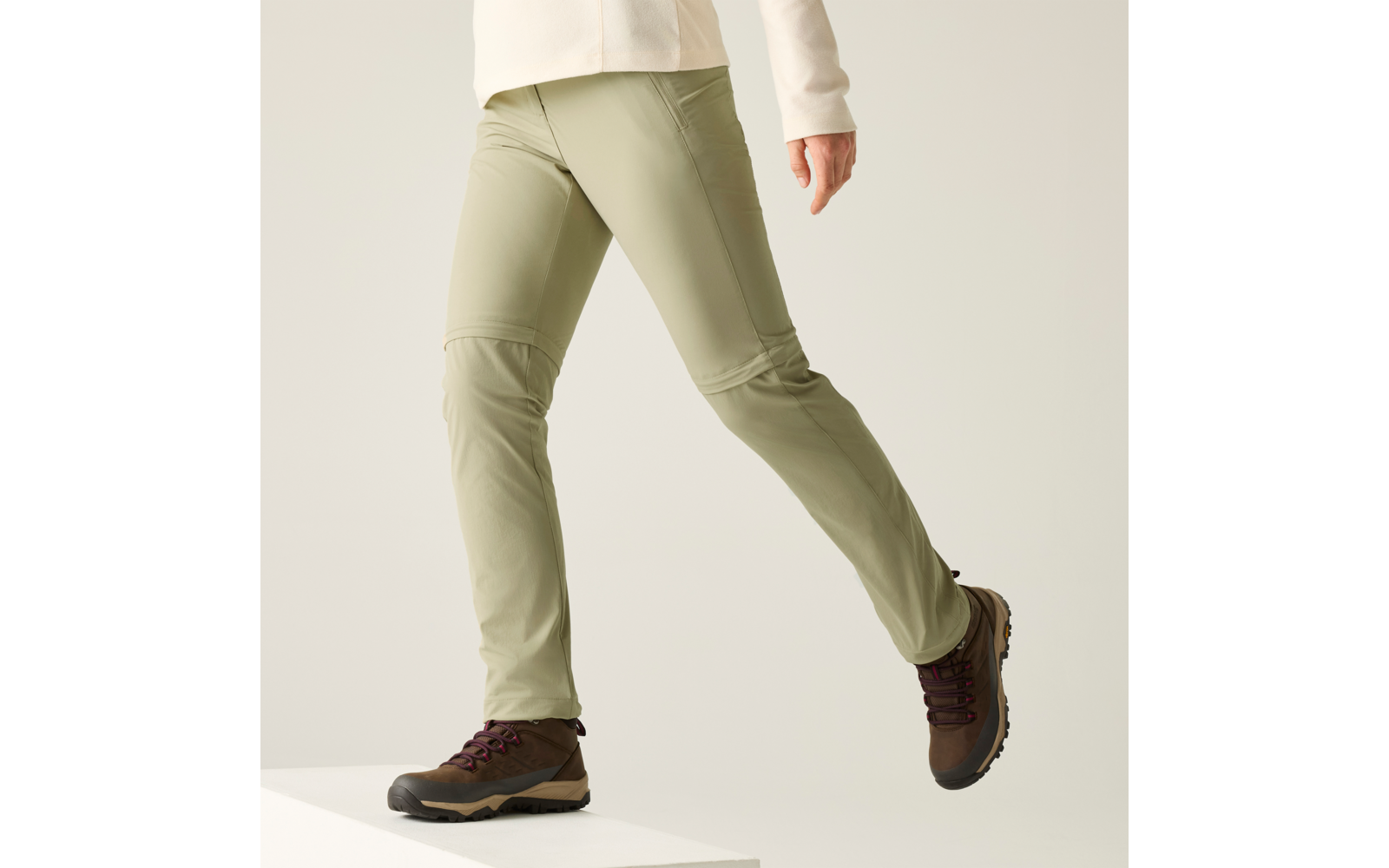 Beinpartie einer Person, die von einem weißen Podest steigt, hellgrüne Hose, cremefarbener Ärmel sichtbar, braune Schnürschuhe