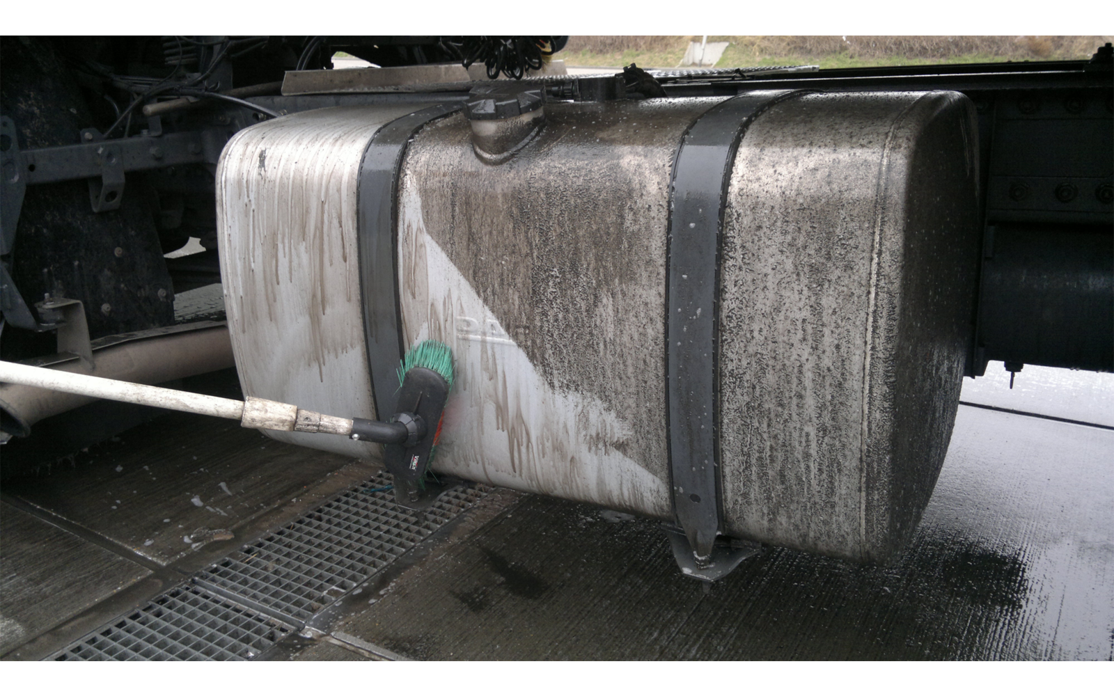 Metalltank unter einem Lkw, eine grüne Bürste an einer Stange berührt den Tank; nasse Streifen, Metallbänder und Fahrgestell sichtbar