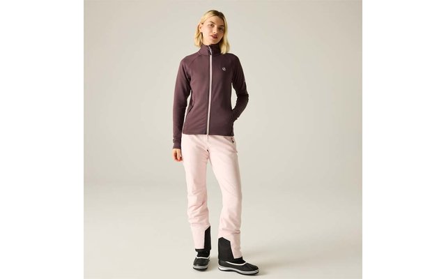 Person mit blonden Haaren steht vor hellem Hintergrund, trägt dunkelviolette Reißverschlussjacke, hellrosa Hosen und schwarze Stiefel, eine Hand in einer Tasche