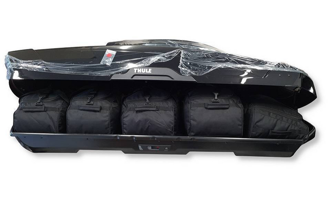 Coffre de toit noir ouvert avec logo Thule, contenant quatre sacs de voyage noirs côte à côte, partiellement recouverts d'un film plastique transparent