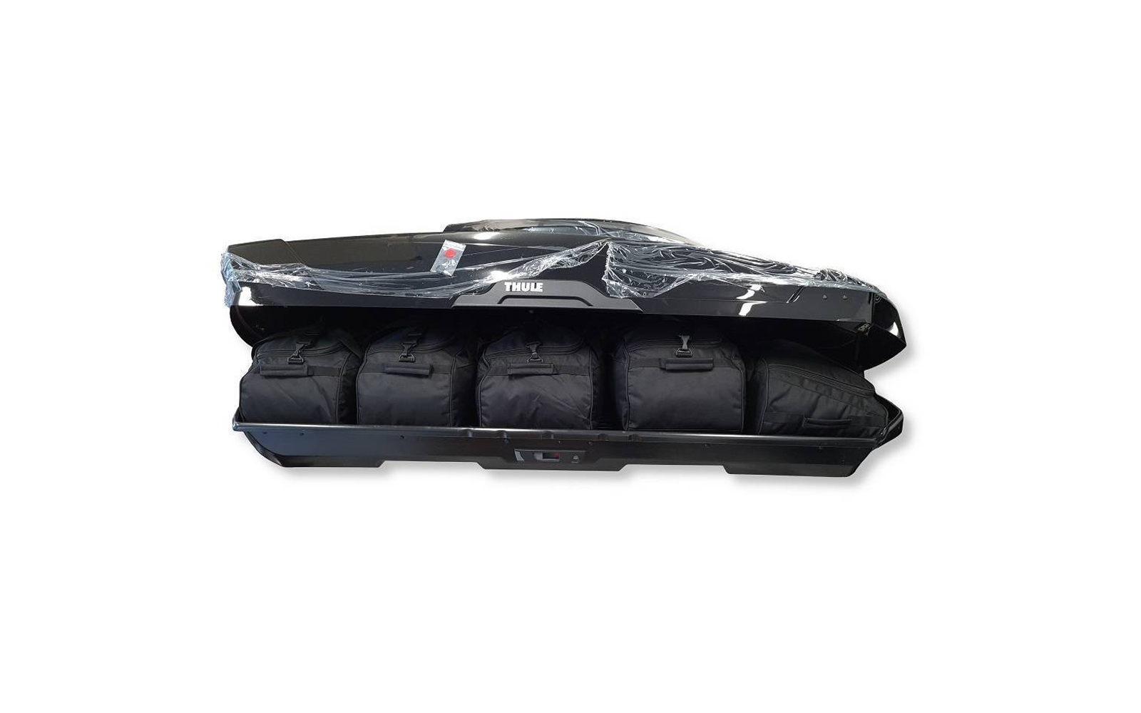 Coffre de toit noir ouvert avec logo Thule, contenant quatre sacs de voyage noirs côte à côte, partiellement recouverts d'un film plastique transparent