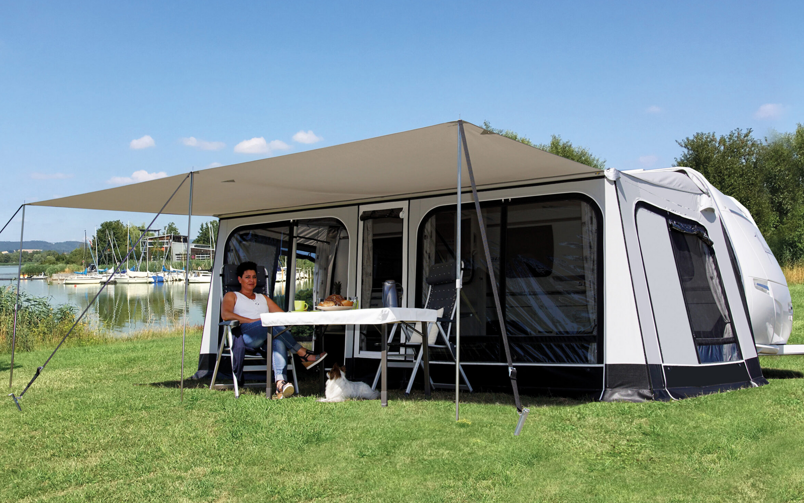 Wohnwagen mit Vorzelt auf einer Wiese am See; unter dem Vorzelt sitzt eine Person an einem Tisch, ein kleiner Hund liegt unter dem Tisch; im Hintergrund Wasser, Boote und Bäume