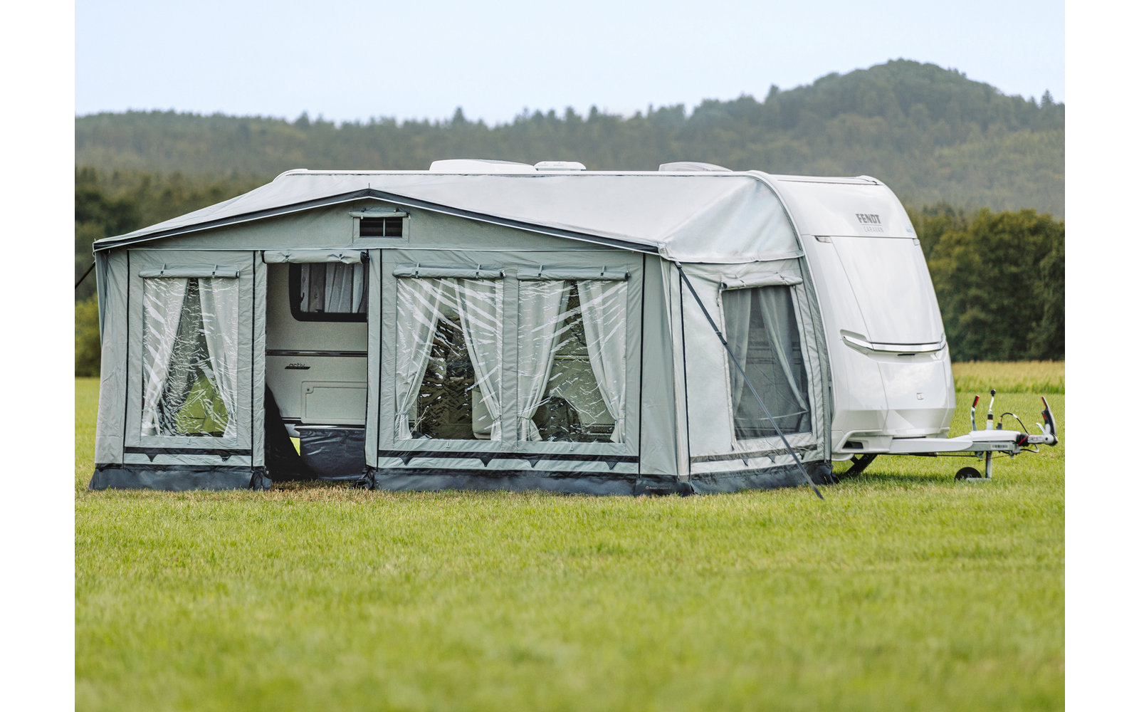 Caravane avec auvent fixé sur un champ herbeux, arbres et basses collines en arrière‑plan