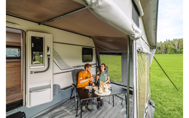 Zwei Personen sitzen an einem Tisch unter einem Vorzelt an einem Wohnwagen auf einer Wiese; auf dem Tisch stehen Teller, Tassen und Flaschen