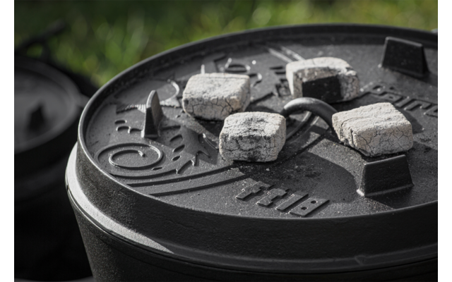 Gusseiserner Topfdeckel mit Kohlebriketts und Ascheresten, unscharfer Rasenhintergrund