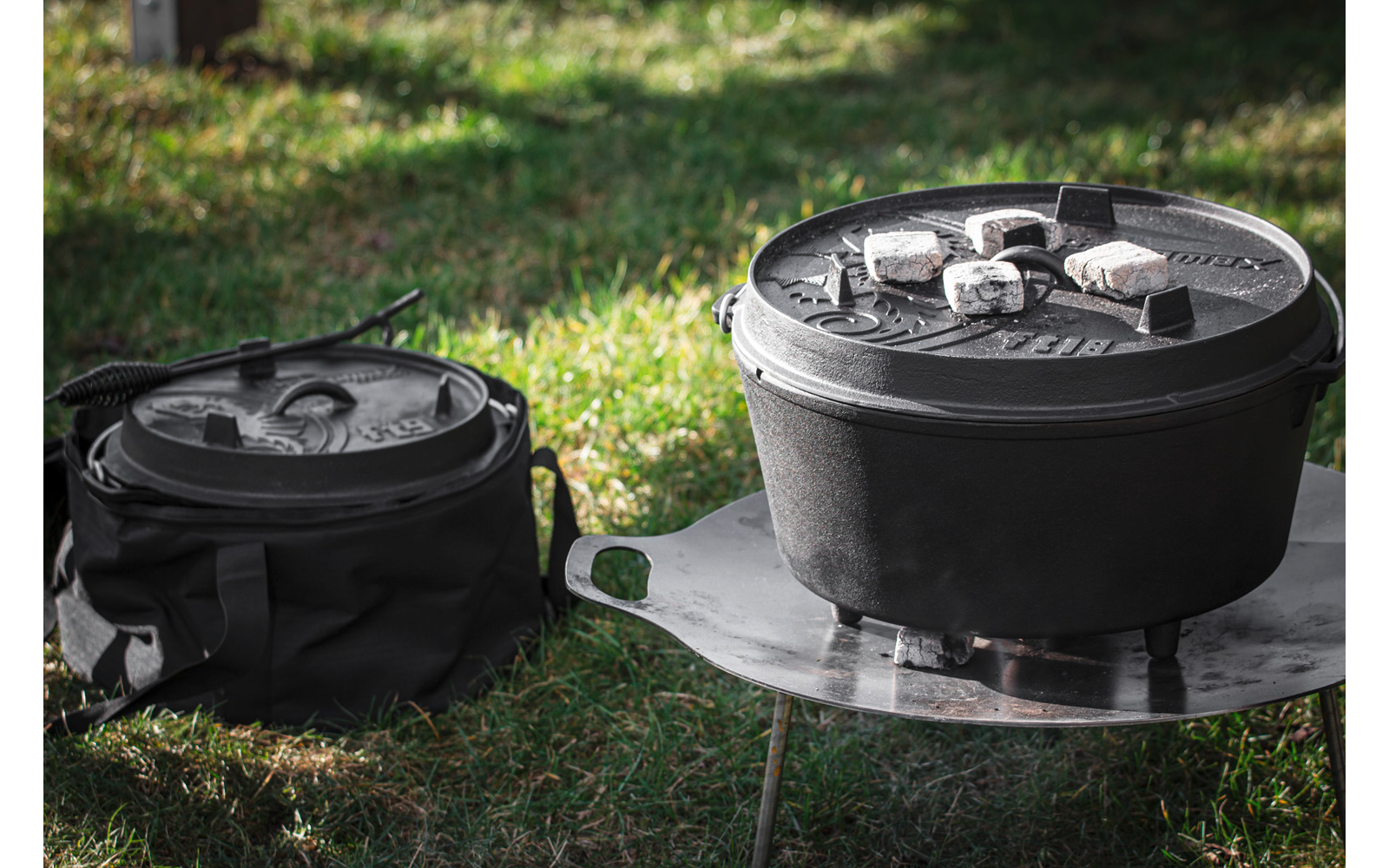 Zwei gusseiserne Töpfe auf Gras; rechter Topf auf runder Metallfeuerstelle, mit Kohlebriketts auf dem Deckel; linker Topf in schwarzer Tragetasche mit Deckel