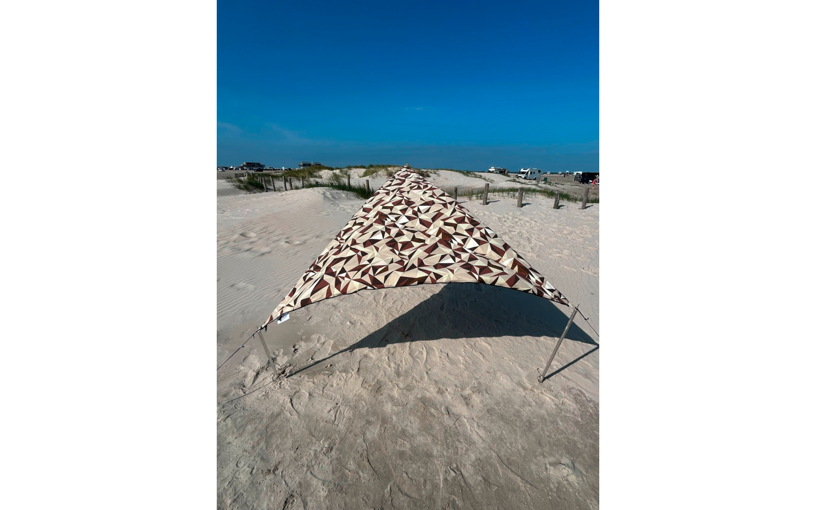 Dreieckiges Sonnensegel mit braun-beigem Muster auf sandigem Strand, Metallstangen, Dünen mit Strandgras und blauem Himmel im Hintergrund