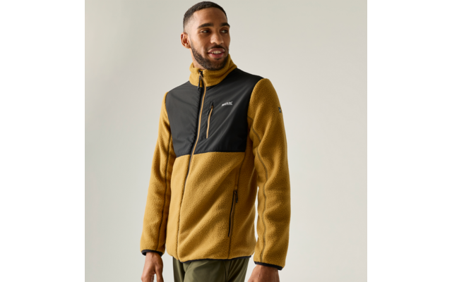 Person, stehend, blickt zur Seite, trägt senfgelbe Fleecejacke mit schwarzem Brustbereich und Reißverschlusstasche, olivgrüne Hose, heller einfarbiger Hintergrund