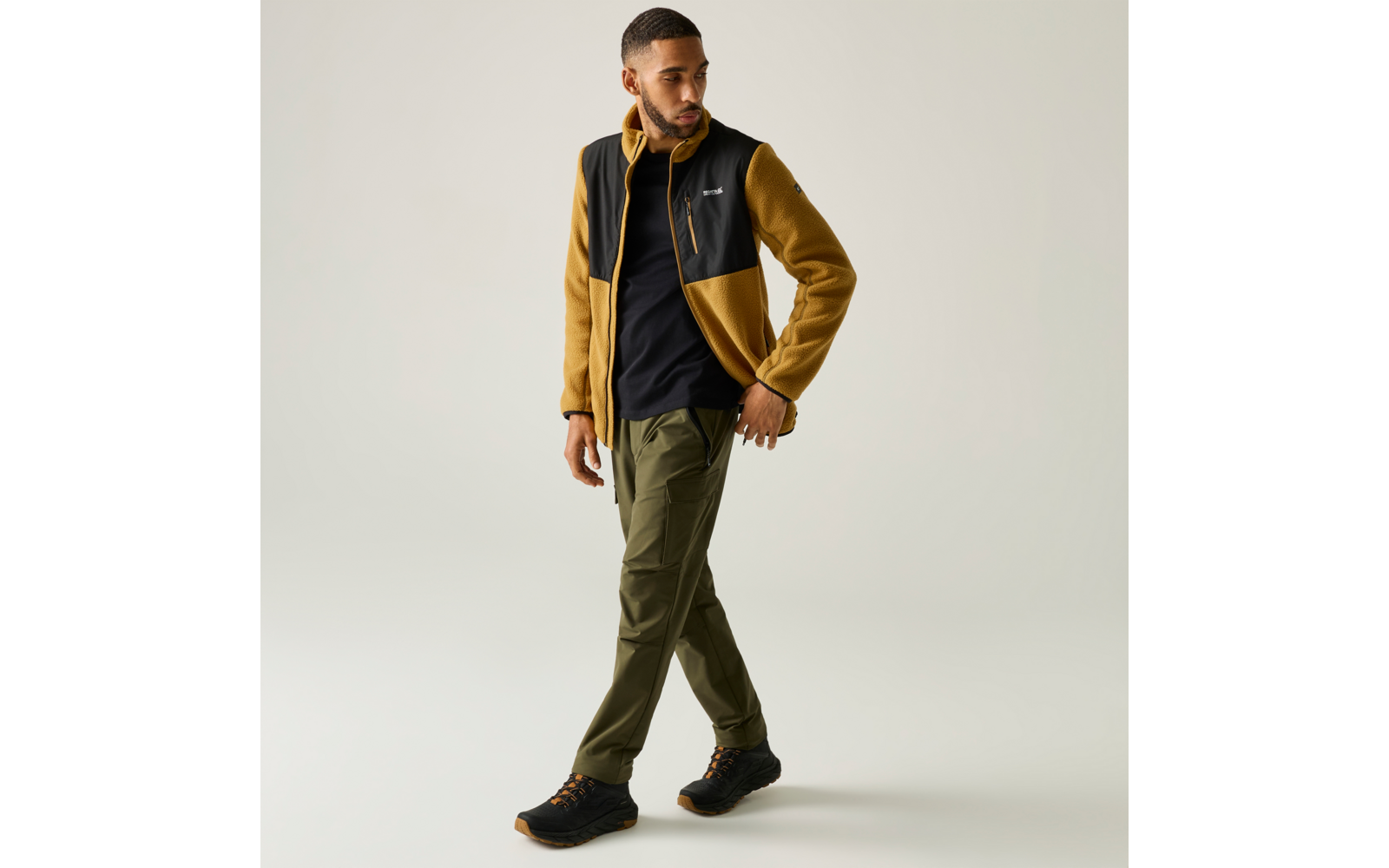 Mann in gelb-schwarzer Fleecejacke, schwarzem T-Shirt, olivgrünen Hosen und schwarzen Wanderschuhen, geht vor hellem Hintergrund, Blick nach links