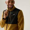 Person trägt senfgelbe Fleecejacke mit schwarzer Brustpartie, hält Reißverschluss, lächelt, heller Hintergrund