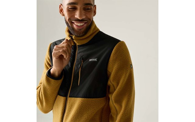 Person trägt senfgelbe Fleecejacke mit schwarzer Brustpartie, hält Reißverschluss, lächelt, heller Hintergrund