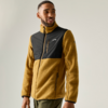 Person, stehend, blickt zur Seite, trägt senfgelbe Fleecejacke mit schwarzem Brustbereich und Reißverschlusstasche, olivgrüne Hose, heller einfarbiger Hintergrund
