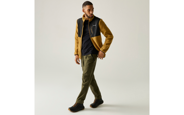 Mann in gelb-schwarzer Fleecejacke, schwarzem T-Shirt, olivgrünen Hosen und schwarzen Wanderschuhen, geht vor hellem Hintergrund, Blick nach links