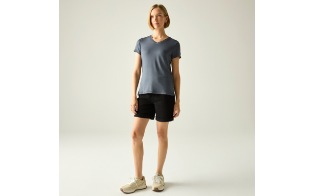 Stehende Person vor neutralem Hintergrund, kurze blonde Haare, trägt blau-graues V‑T-Shirt, schwarze Shorts und beige Sneaker