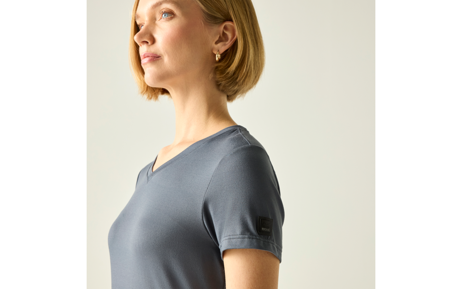 Seitliches Porträt einer Person mit kurzem blondem Bob, goldenen Creolen und graublauem T‑Shirt mit V-Ausschnitt vor hellem Hintergrund