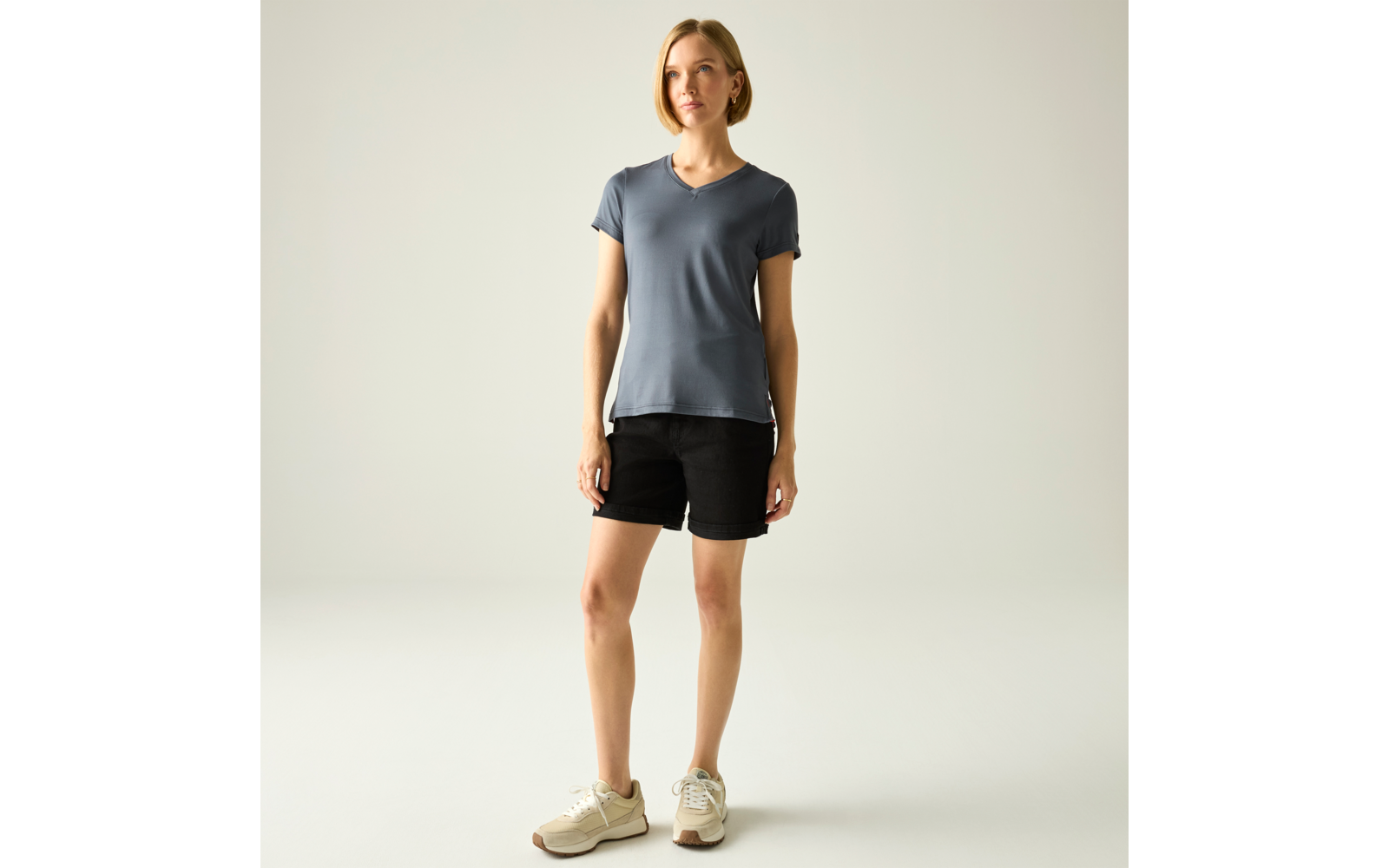 Stehende Person vor neutralem Hintergrund, kurze blonde Haare, trägt blau-graues V‑T-Shirt, schwarze Shorts und beige Sneaker