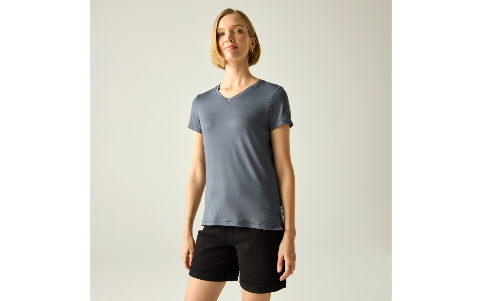 Frau steht vor hellem Hintergrund, trägt graublaues T‑Shirt mit V‑Ausschnitt und schwarze Shorts, Hände an den Seiten