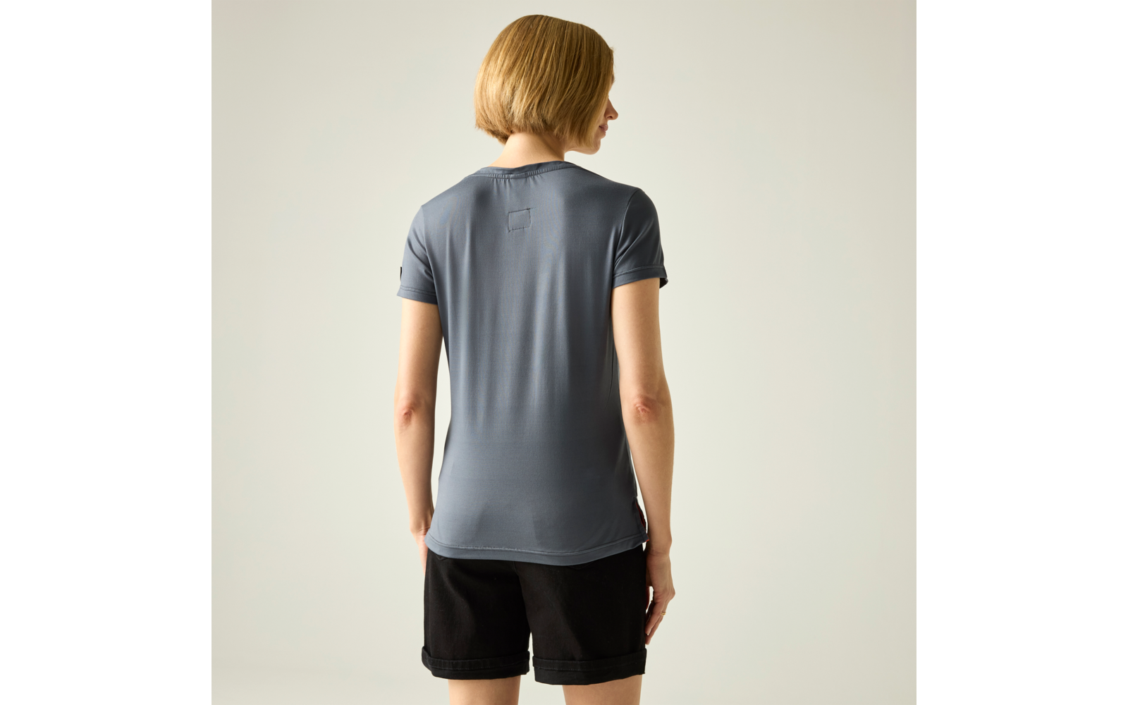 Kurzhaarige Person von hinten, trägt graues T‑Shirt mit kurzen Ärmeln und schwarze Shorts, steht vor hellem einfarbigem Hintergrund