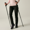 Parte inferiore del corpo di una persona, visibili solo gambe e mani, che tiene due bastoncini da trekking neri; pantaloni neri, maniche rosa chiaro, scarpe da trekking viola su sfondo chiaro