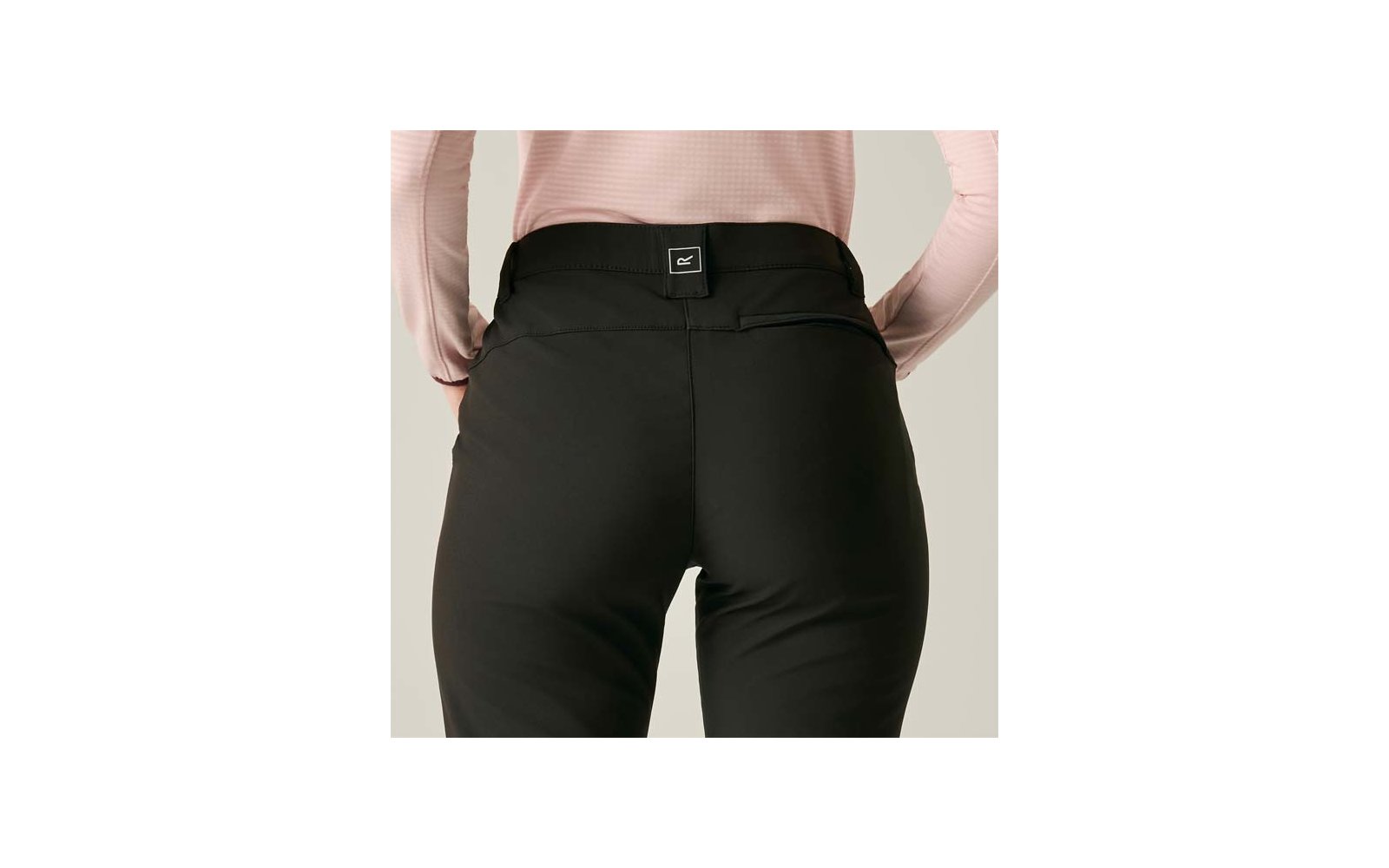 Achteraanzicht van een persoon van taille tot bovenbenen, zwarte broek, lichtroze top met lange mouwen in de taille gestopt, klein vierkant label in het midden van de tailleband, neutrale achtergrond