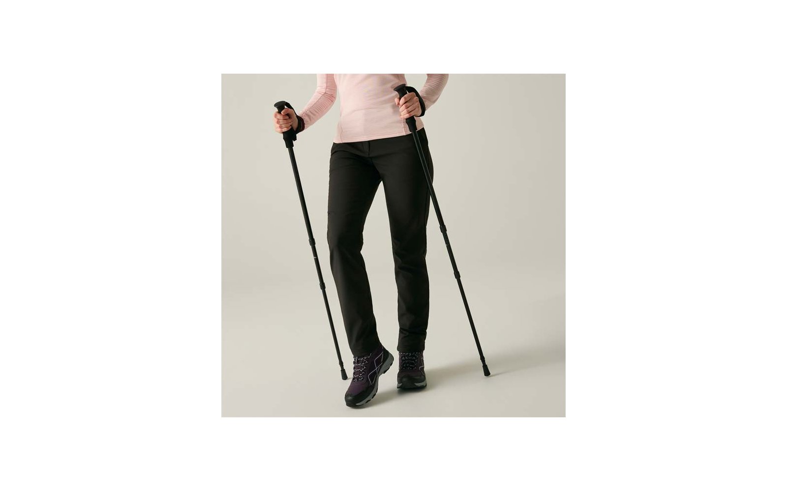 Onderlichaam van een persoon, alleen benen en handen zichtbaar, houdt twee zwarte wandelstokken vast; zwarte broek, lichtroze mouwen, paarse wandelschoenen op een lichte achtergrond