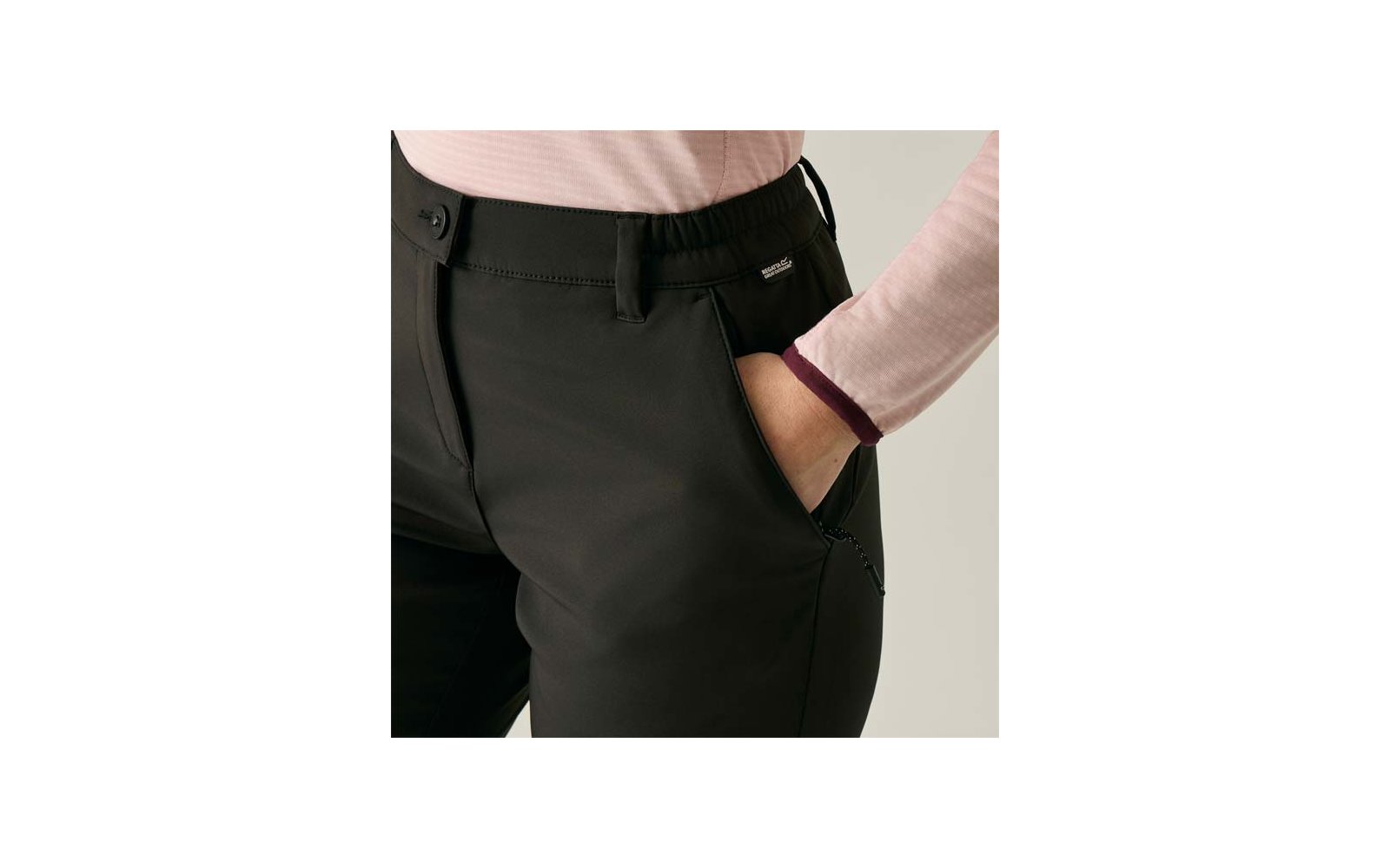 Onderlichaam van een persoon in zwarte broek, rechterhand in de voorzak, lichtroze top met lange mouwen en donker boord, zichtbare riemlussen en klein logo bij de zak