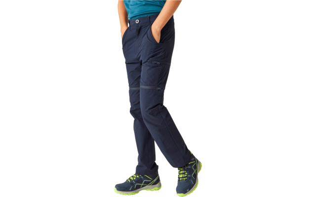 Lower body of a person wearing dark blue pants with side pockets, hands in the pockets, a turquoise top partly visible, blue shoes with neon green soles