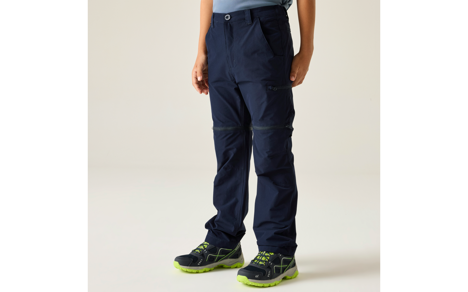 Lower body of a person wearing dark blue pants with a zipper at the knee, side pocket and blue waistband button; dark athletic shoes with neon green soles; light background