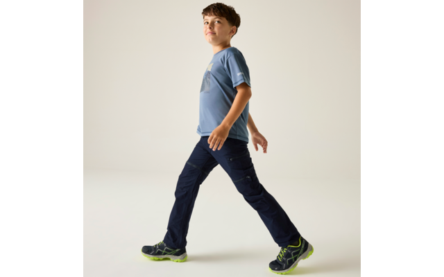 A boy in a blue T-shirt and dark pants walks to the right on a light background, wearing dark sneakers with green soles