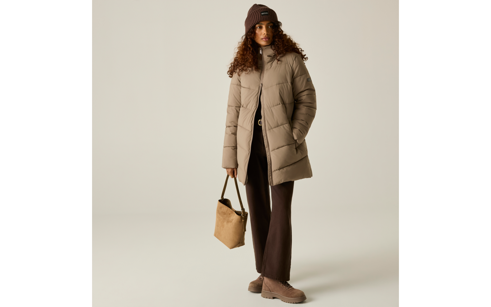 Persona con capelli castani ricci, berretto di maglia marrone, giacca imbottita lunga beige, pantaloni scuri, tiene una borsa beige, in piedi su sfondo chiaro