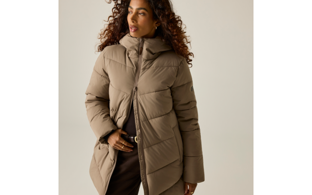 Persona con capelli ricci che indossa un piumino beige trapuntato con cappuccio, cerniera parzialmente aperta; una mano vicino a pantaloni con fibbia; sfondo chiaro