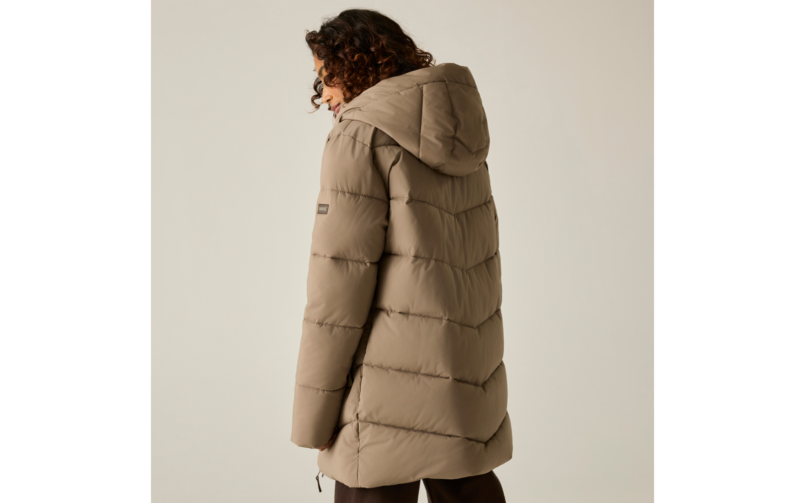 Persona vista da dietro con capelli ricci scuri, indossa un cappotto imbottito trapuntato beige con cappuccio e una toppa logo visibile sulla manica, pantaloni scuri, sfondo chiaro uniforme