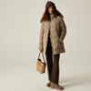 Persona con capelli castani ricci, berretto di maglia marrone, giacca imbottita lunga beige, pantaloni scuri, tiene una borsa beige, in piedi su sfondo chiaro