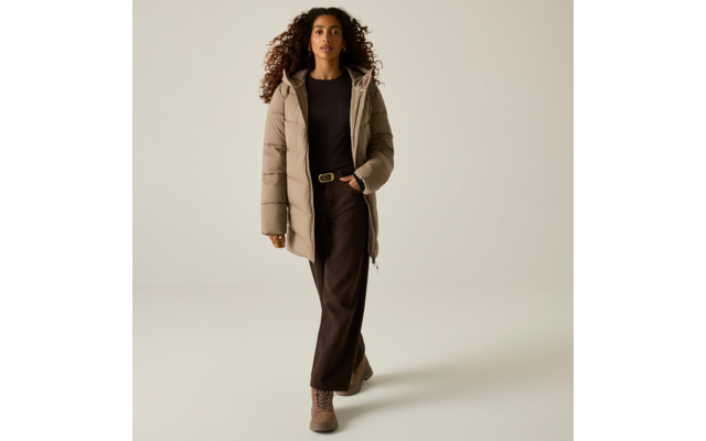Persona con capelli lunghi e ricci che indossa un cappotto trapuntato beige, una maglia scura, pantaloni scuri e stivaletti marroni, che cammina su sfondo chiaro