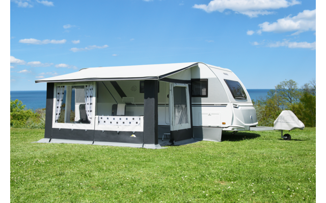 Weißer Wohnwagen mit angebautem dunkelgrauen Vorzelt auf grünem Rasen, Meer im Hintergrund, blauer Himmel mit vereinzelten Wolken, kleines weißes Zelt rechts vom Wohnwagen