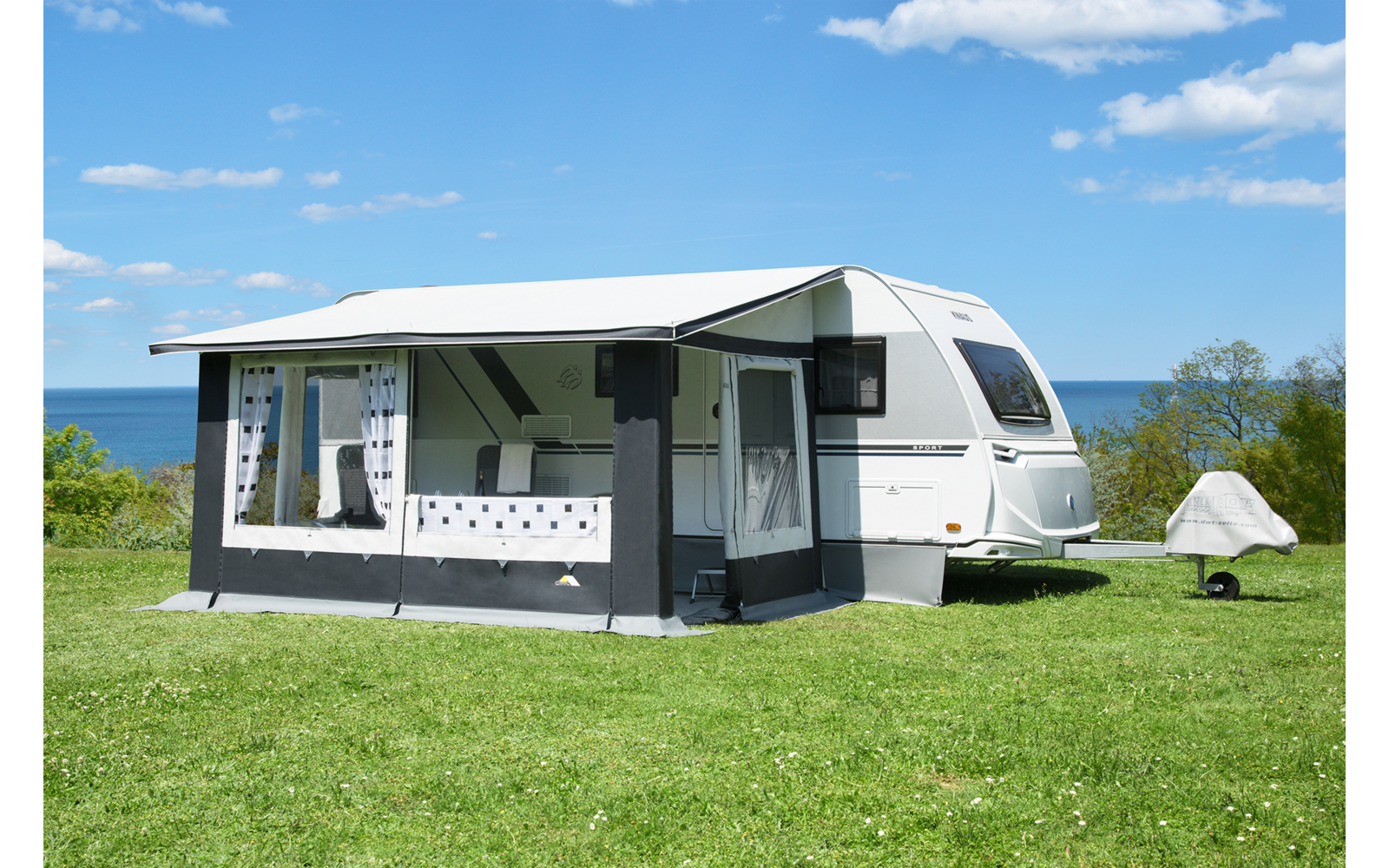 Weißer Wohnwagen mit angebautem dunkelgrauen Vorzelt auf grünem Rasen, Meer im Hintergrund, blauer Himmel mit vereinzelten Wolken, kleines weißes Zelt rechts vom Wohnwagen