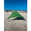 Dreieckiges, grün gemustertes Tuch auf Sand, gehalten von Metallstangen; im Hintergrund Dünen, Holzzaun, einige Gebäude und blauer Himmel