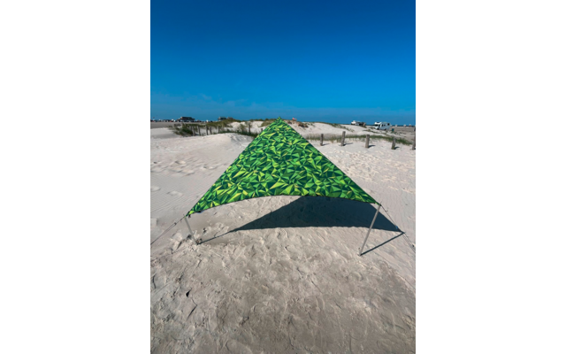 Dreieckiges, grün gemustertes Tuch auf Sand, gehalten von Metallstangen; im Hintergrund Dünen, Holzzaun, einige Gebäude und blauer Himmel