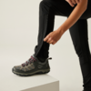 Person zieht eine schwarze Socke über den linken Fuß, trägt schwarze Hose und graue Sportschuhe