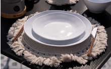 Plato blanco sobre una mesa con un mantel individual gris, al lado una taza blanca y cubiertos