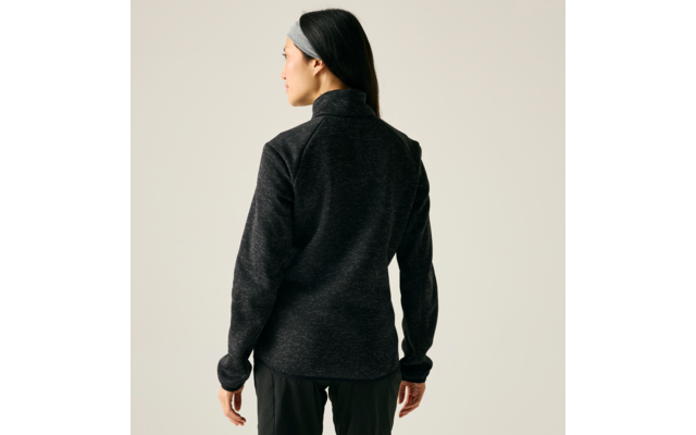 Person von hinten, trägt dunkelgraue Jacke mit Stehkragen, schwarzes Haar mit grauem Stirnband, schwarze Hose, heller neutraler Hintergrund