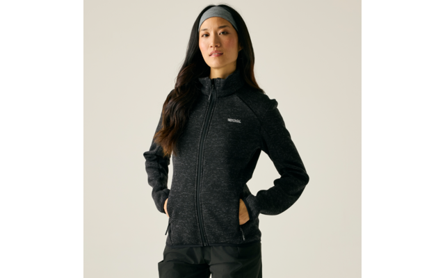 Person steht vor hellem Hintergrund, trägt dunkle Fleecejacke mit durchgehendem Reißverschluss und kleinem Logo, Hände in Seitentaschen, Stirnband, lange dunkle Haare