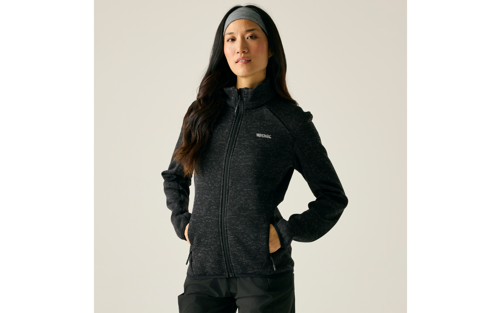 Person steht vor hellem Hintergrund, trägt dunkle Fleecejacke mit durchgehendem Reißverschluss und kleinem Logo, Hände in Seitentaschen, Stirnband, lange dunkle Haare