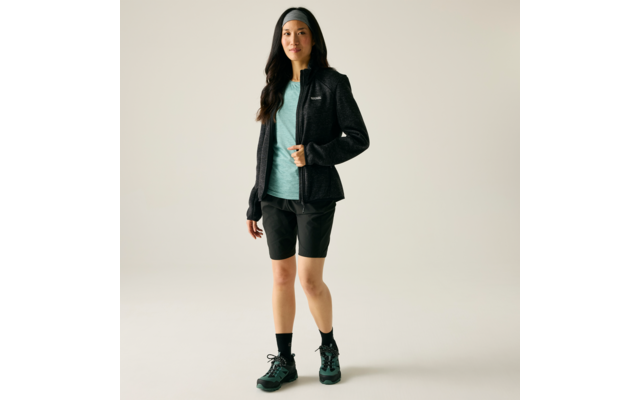 Person steht auf hellem Hintergrund, trägt schwarzes Stirnband, schwarze Jacke mit Reißverschluss, türkisfarbenes T-Shirt, knielange schwarze Shorts, schwarze Socken und dunkelgrüne Wanderschuhe