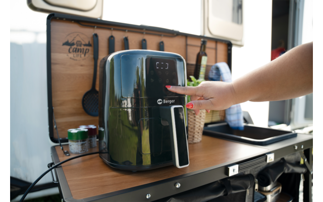 Friteuse à air noire sur un plan de travail en bois rabattable d’un camping-car ; main appuie sur le panneau de commande ; bouteilles, boîtes et ustensiles en arrière-plan