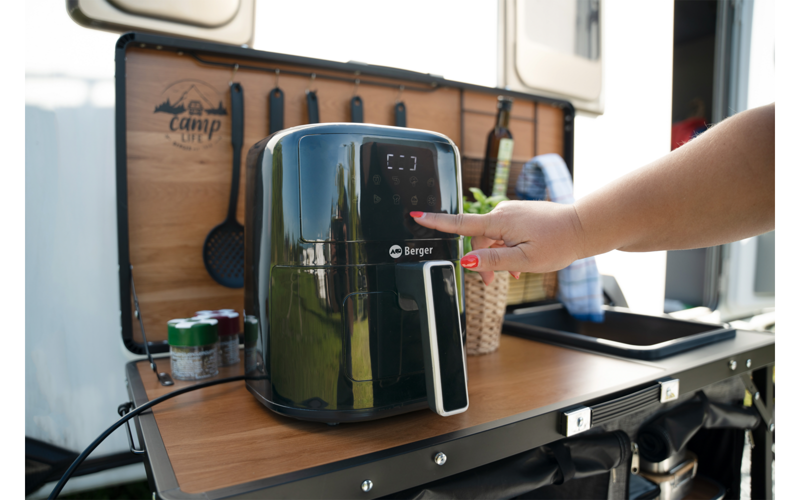 Friteuse à air noire sur un plan de travail en bois rabattable d’un camping-car ; main appuie sur le panneau de commande ; bouteilles, boîtes et ustensiles en arrière-plan