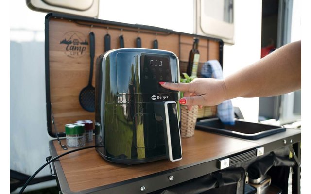 Schwarze Heißluftfritteuse auf klappbarer Holzarbeitsfläche eines Campers; Hand drückt Bedienfeld; Flaschen, Dosen und Küchenutensilien im Hintergrund