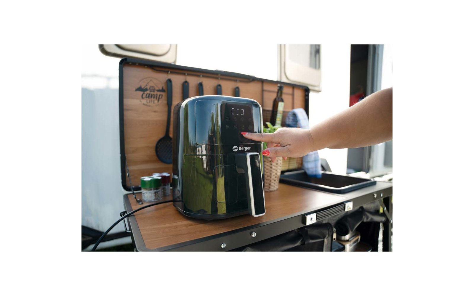 Schwarze Heißluftfritteuse auf klappbarer Holzarbeitsfläche eines Campers; Hand drückt Bedienfeld; Flaschen, Dosen und Küchenutensilien im Hintergrund