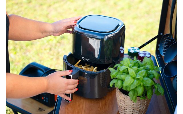 Zwei Hände öffnen die Schublade einer schwarzen Heißluftfritteuse mit Pommes; Fritteuse steht auf einer Holzfläche, daneben ein Topf mit Basilikum und Gewürzgläser; grüner Rasen im Hintergrund